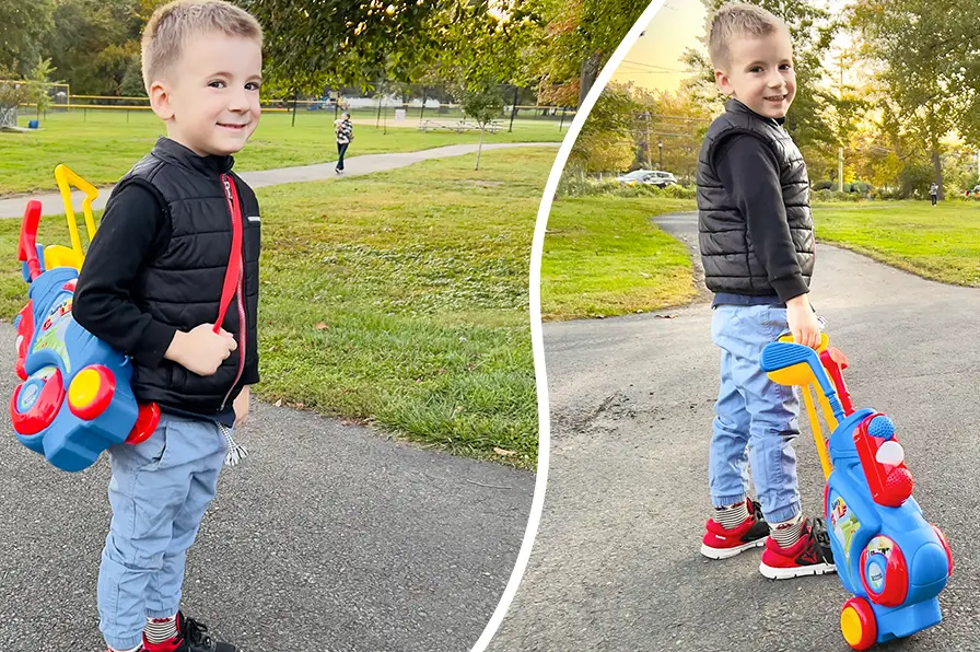 A toddler carrying and pulling his toy golf club set through a park.