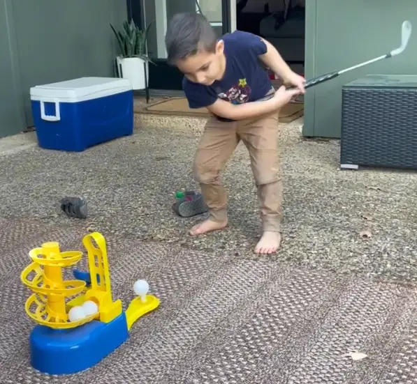 A young child swinging a golf club at a ball on the tee of a golf trainer for kids.