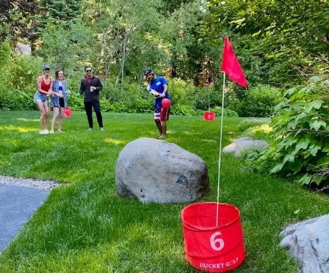 A group of people playing BucketGolf in a backyard, hitting golf balls towards buckets (holes).