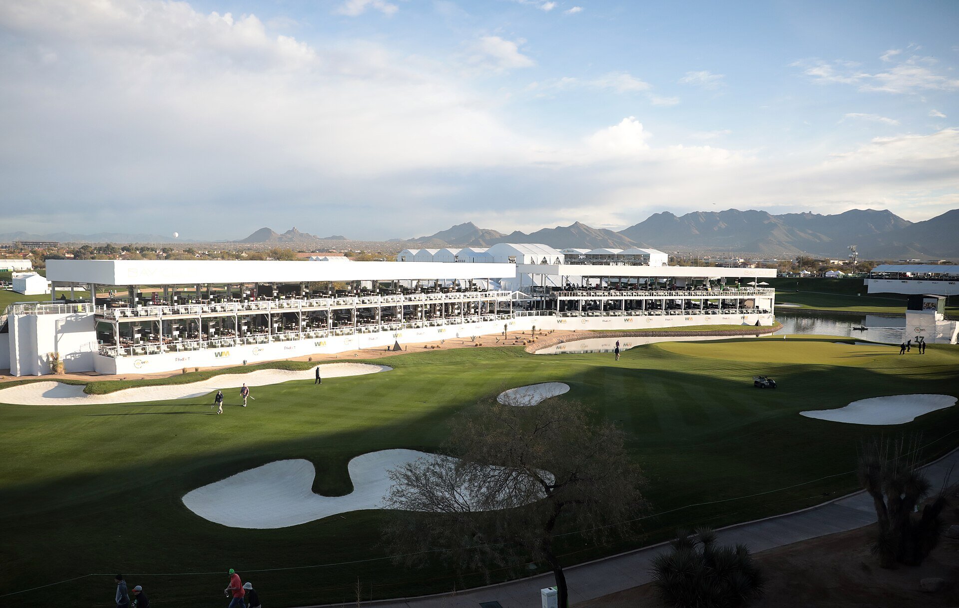 TPC Scottsdale Stadium Course during the 2020 Waste Management Phoenix Open, with grandstands around the hole, ryegrass fairways, bunkers, and the McDowell Mountains beyond.