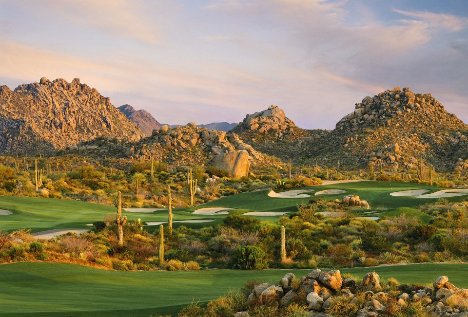 A desert golf hole at Troon North Golf Club in Scottsdale, Arizona, with a green and bunkers set among granite boulders, saguaros, and Pinnacle Peak at sunset.